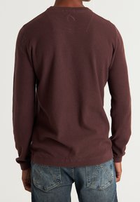 Pull en maille marron avec un motif texturé, manches longues et ourlet côtelé, associé à un jean bleu délavé visible à la taille.