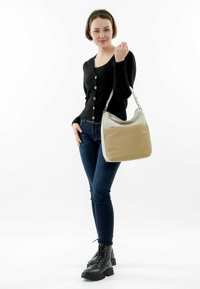 Bolso ligero en beige y crema, con una textura suave y una correa plateada. La modelo lleva un cárdigan negro y jeans oscuros.