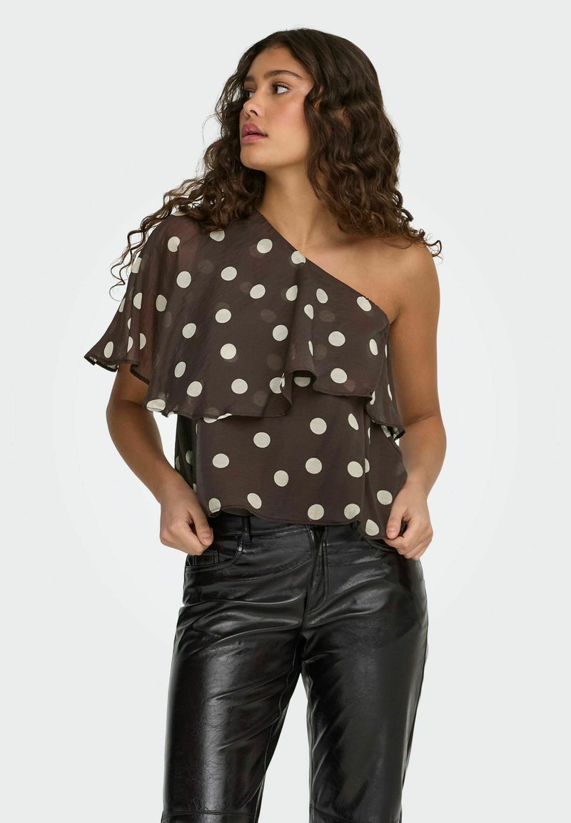 Femme portant un haut brun à pois avec un épaulière et des volants, associé à un pantalon en cuir noir, regardant vers sa gauche.