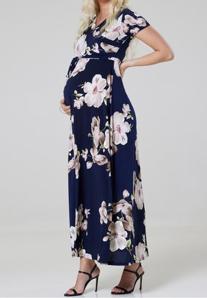 Femme enceinte en robe longue fleurie bleu marine et sandales à talons noires, main sur la hanche et soutenant le ventre, debout devant un fond uni.