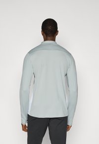 Ανοιχτό γκρι μακρυμάνικο πουκάμισο με φερμουάρ στον γιακά, με λευκές πλαϊνές πανέλες σε αντίθεση και λεία, ελαστική υφή υφάσματος.