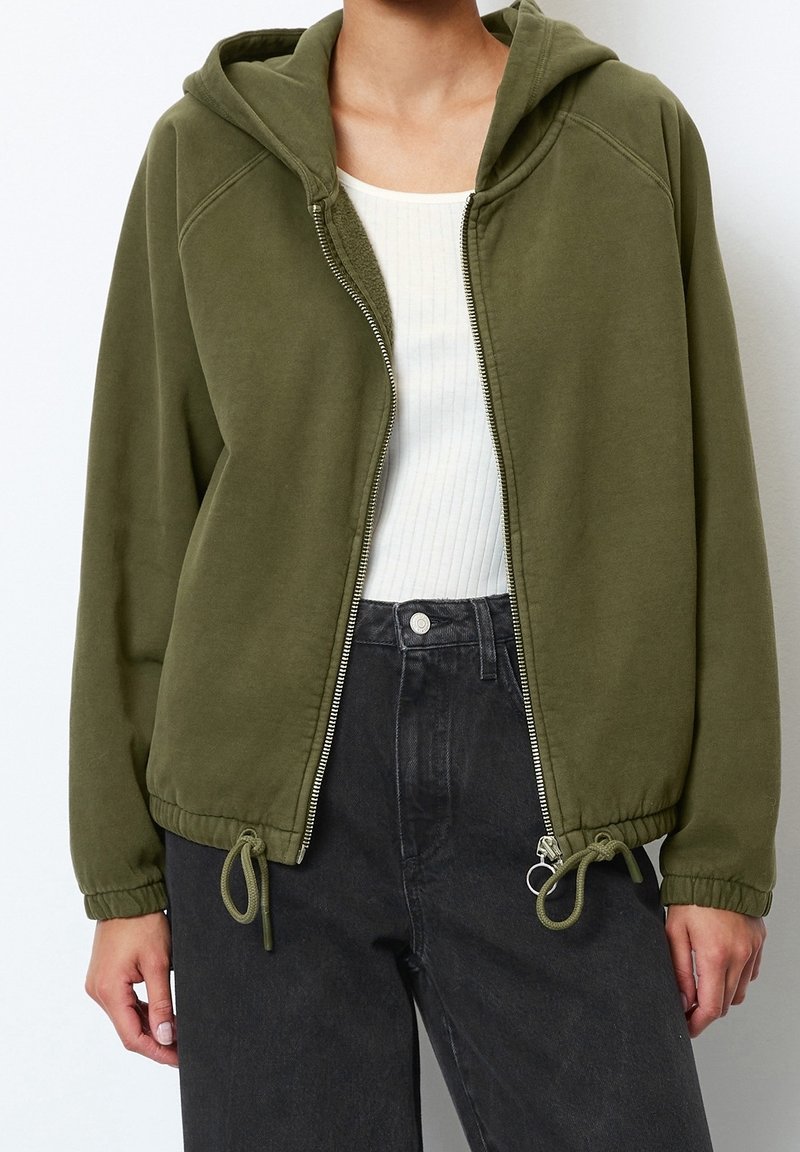 Felpa con zip color verde oliva, con un bordo regolabile, realizzata in un tessuto leggero e morbido e dotata di una cerniera frontale. Indossata sopra una maglietta a coste bianca.
