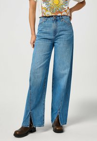 Jeans anchos de color azul claro con cintura alta, bolsillos frontales y aberturas laterales, combinados con botas de tobillo marrones. Textura de denim de algodón.