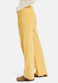 Pantalons en velours côtelé jaunes à jambes larges avec des côtes verticales, deux poches arrière et une taille haute, associés à des chaussures marron.