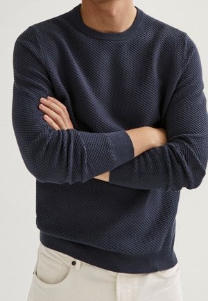Osoba nosząca ciemny, teksturowany sweter z długim rękawem, z założonymi skrzyżowanymi rękami, w połączeniu z jasnymi spodniami.