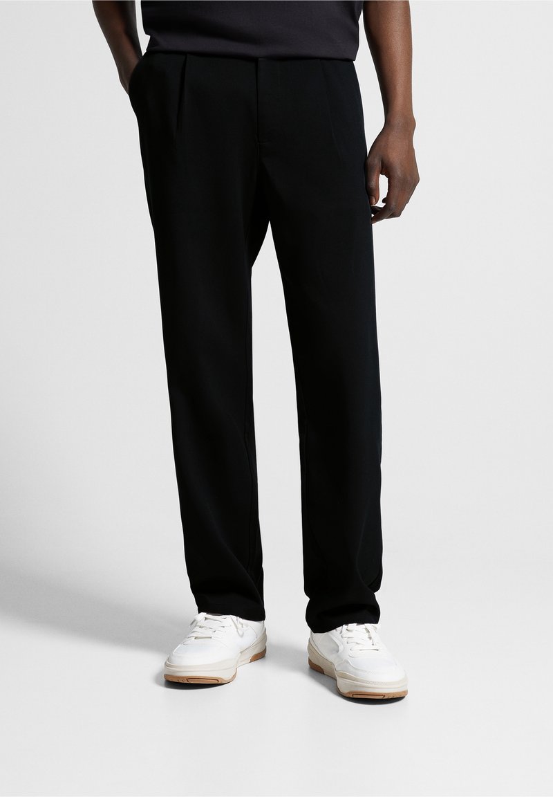 Homme portant un pantalon noir à jambes droites et des baskets blanches, debout avec une main dans la poche, devant un fond uni.