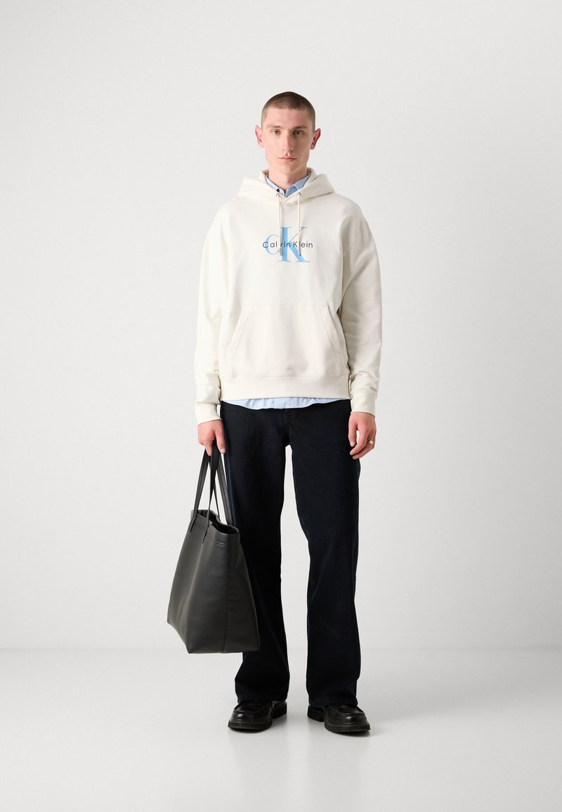 Felpa crema con logo blu, indossata sopra una camicia button-up azzurra chiara. Jeans scuri e borsa a tracolla in pelle nera. Scarpe nere. Design semplice.