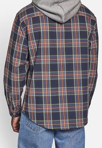 Tył koszuli z długim rękawem w kratę w kolorze granatowym z pomarańczowymi i żółtymi liniami, z zaokrąglonym dołem i mankietami na guziki, noszona na szarej bluzie z kapturem.