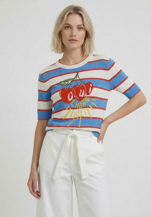 Blonde vrouw draagt een blauw-wit gestreept topje met rode kersen en de tekst "CHEER", gecombineerd met een witte broek met hoge taille, gestrikt in de taille.