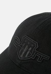 Čierna čiapka vyrobená z látky s vystúpeným čierne vyšitou logom s hviezdami a pruhmi vpredu. Zakrivený šilt a textúrovaný povrch.