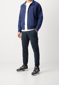 Marineblauwe bomberjack met een rits aan de voorkant en geribde manchetten, gecombineerd met donkere cargobroeken en New Balance sneakers met kleurrijke accenten.