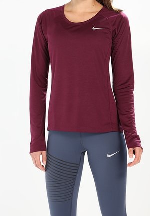 Kobieta ubrana w długi rękaw burgundowy top sportowy oraz granatowe legginsy z czarnymi paskami i logo Nike na klatce piersiowej i udzie.