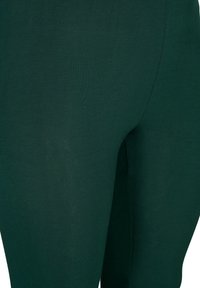 Close-up van donkergroene leggings gemaakt van gladde, lichte stof. Heeft een aansluitend ontwerp met een subtiele glans, zonder patronen of accenten.