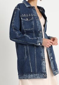 Jeansová bunda v tmavě modré barvě s využitím stříbrných hrotů a ozdob. Má límec, přední kapsy a zapínání na knoflíky. Viditelná struktura kamenného obarvení.
