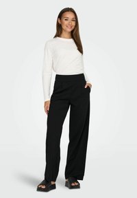 Sorte wide-leg bukser med høj talje og forlommer, parret med en langærmet hvid bluse. Modellen står foran en ensfarvet baggrund.