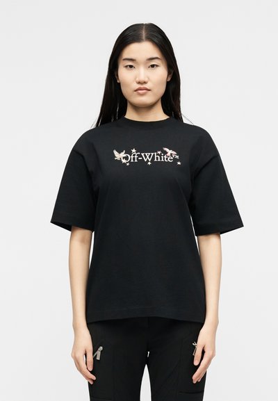 Νέα γυναίκα με μακριά σκούρα μαλλιά που φοράει μαύρο t-shirt Off-White με σχέδιο πουλιού και αστέρι, στέκεται μπροστά σε λευκό φόντο.