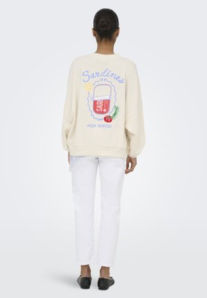Frau mit Dutt, trägt einen cremefarbenen Sweatshirt mit buntem Sardinendosen-Motiv und weißen Hosen, steht vor einfachem Hintergrund.