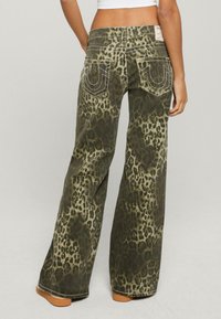 Zielone dżinsy w panterkę z dzwonami, z kontrastowymi stębnowaniami, dużymi tylnymi kieszeniami i luźnym krojem. Tkanina wydaje się mieć fakturę.