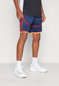 Marine sports shorts med røde og gule detaljer, som har en mesh-struktur, sidelægger og en split-nederdel-design, parret med hvide sneakers.