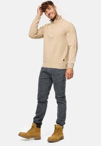 Pull en tricot beige avec col montant et trois boutons, associé à un pantalon gris slim-fit et des bottes à lacets beige.