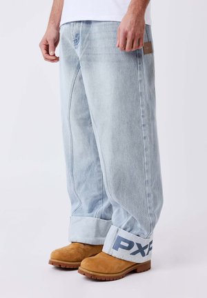 Jean large bleu clair avec de grandes revers affichant le texte "PXF", porté avec des bottines en daim marron clair et une chemise blanche partiellement visible au-dessus de la taille.