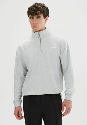 Hombre con sudadera gris claro con cremallera hasta un cuarto y pantalones negros, de pie frente a un fondo blanco liso.