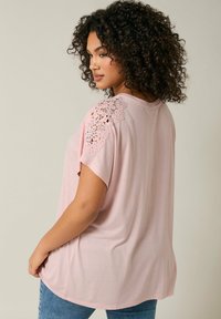 Ljusrosa t-shirt med korta ärmar, med spetsdetaljer på axlarna, avslappnad passform och rundad nederkant. Mjuk tygkänsla.