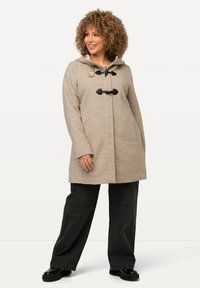 Cappotto beige in misto lana con cappuccio, chiusure a toggle e maniche lunghe. Abbinato a pantaloni neri a gamba larga e scarpe nere robuste.