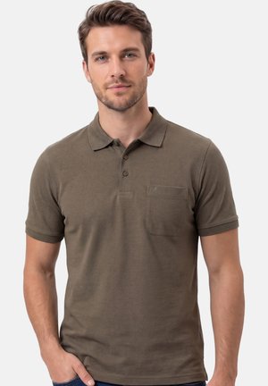 Homme aux cheveux bruns courts portant un polo à manches courtes vert olive avec une poche poitrine, debout devant un fond clair uni.