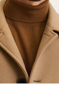 Bruine wollen jas met een nekkraag met inkeping, voorzien van een knoopsluiting en zijzakken. Daaronder is een geribbelde bruine coltrui zichtbaar.