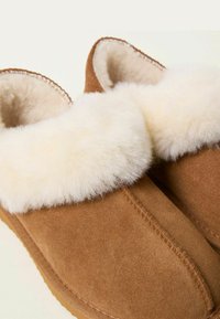 Brown suede slippers featuring a fluffy white fur cuff, rounded design, contrasting stitching, and a flexible brown rubber sole.