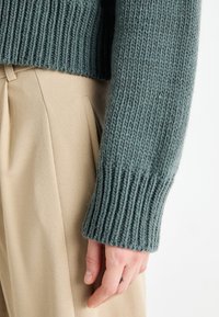 Ribbed knit sweater in muted green, featuring a straight cuff. Paired with beige pleated trousers, highlighting fabric texture and structure.