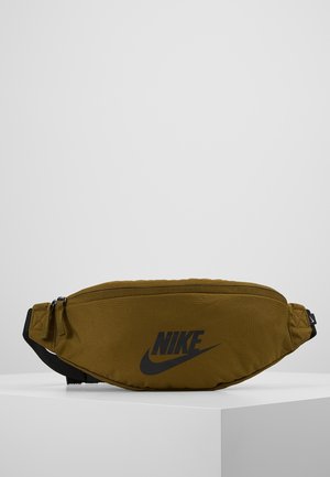 Riñonera Nike verde oliva con logotipo y cremallera negros, expuesta sobre una superficie blanca y un fondo claro y sencillo. Sin personas presentes.
