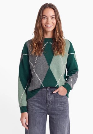 Ung kvinde med langt brunt hår iført en grøn og grå argyle sweater og grå jeans, smilende med den ene hånd i lommen.