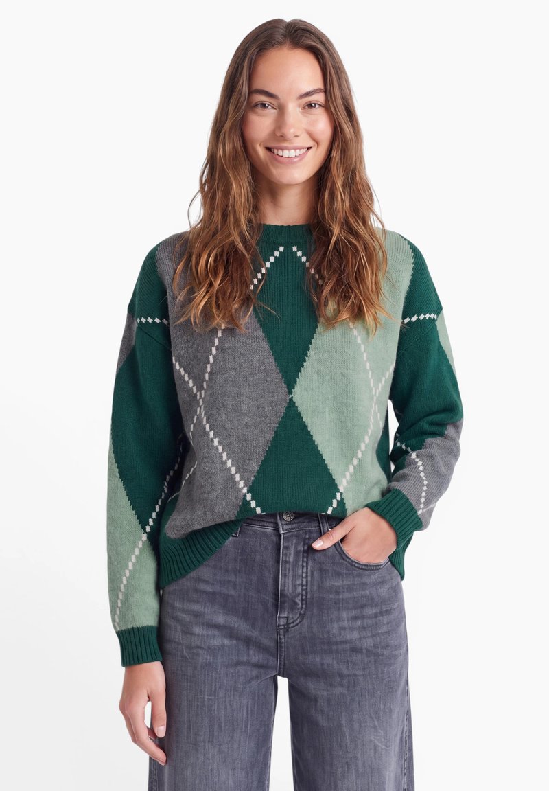 Jeune femme aux longs cheveux bruns portant un pull à losanges vert et gris et un jean gris, souriante, une main dans la poche.
