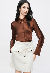 Chemise marron en satin boutonnée avec col, associée à une jupe blanche dotée de poches avant et de boutons dorés. Texture lisse, coupe ajustée.