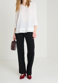 Valkoinen paita, jossa on pleating-yksityiskohtia, yhdistettynä mustiin leveälahkeisiin housuihin. Punaiset korkokengät ja pieni ruskea käsilaukku viimeistelevät asun.