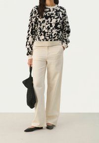 Femme portant un pull à motifs noir et blanc, pantalon beige à jambes larges, ballerines noires, tenant un sac à main tissé noir dans une main.