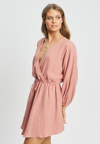 Obleka v breskovo rožnati barvi iz teksturirane tkanine, dolge rokave, elastičen pas in V-izrez. Kratka dolžina z sproščenim krojem.