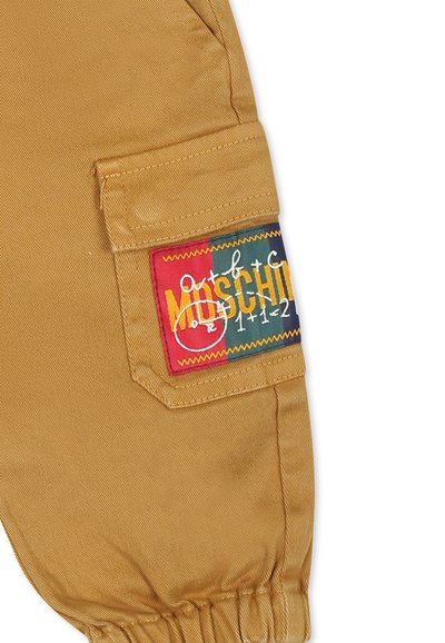 Beige Cargo-Hosen aus Stoff verfügen über eine Seitentasche mit einem bunten, gestickten Etikett, das die Marke und stilisierte Kunstelemente detailliert beschreibt.