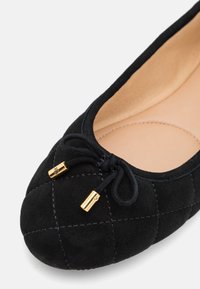 Zwarte suède ballerina met een quiltpatroon, ronde neus en zwarte veters met gouden hardware-accenten. Beige binnenvoering.
