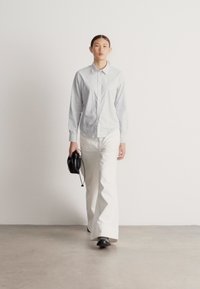 Cămașă cu dungi și nasturi, de culoare albastru deschis, combinată cu pantaloni largi albi. Modelul ține o geantă neagră și poartă pantofi negri.