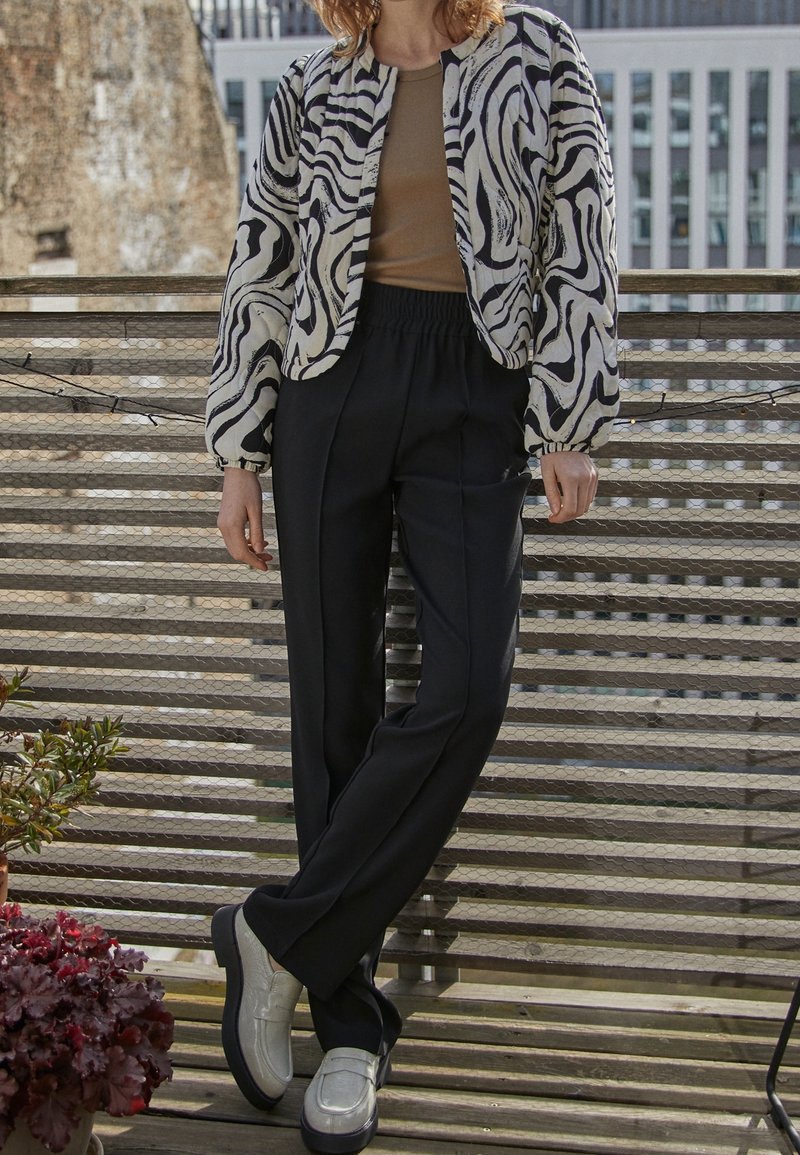 Personne debout sur un toit portant une veste à motif zèbre, une chemise beige, un pantalon noir et des mocassins argentés, avec des plantes à proximité.