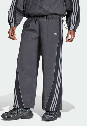 Person trägt dunkle Adidas-Trainingshose und passende Jacke mit schwarzen und grauen Streifen, kombiniert mit schwarzen und grauen Sportschuhen.