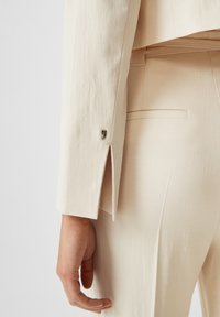 Beige skräddarsydd kavaj i närbild, med en subtil textur, sidoventil och metallisk detalj på manschetten.