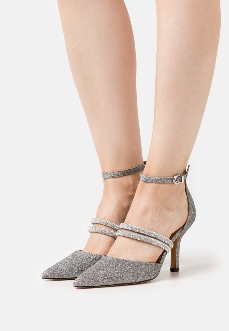 Silver glittrande högklackade skor med spetsiga tår och ankelsnören som har två rader av strass. Smala klackar och slät textur.