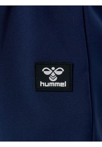 Marineblauwe stof met een gladde textuur heeft een zwart rechthoekig label met het witte "hummel" logo en een gestileerde graphic boven de tekst.