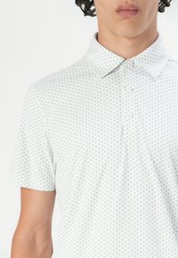 Camisa polo blanca con un pequeño patrón floral azul repetido, un cuello puntiagudo y tres botones en el escote. Mangas cortas.