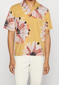 Korte mouwen gele shirt met groot roze en zwart bloemenpatroon, een camp kraag en knoopsluiting aan de voorkant. Draag het over crèmekleurige broek.