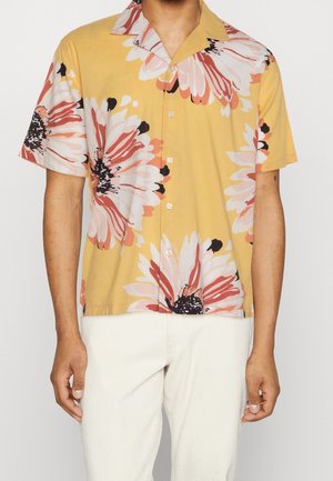 Camisa de manga corta amarilla con un gran patrón floral en rosa y negro, cuello tipo camp y botonadura en la parte frontal. Llevada sobre pantalones de color crema.